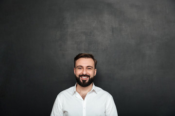 Cropped portrait of young joyful man in white shirt posing on camera with broad smile, isolated over dark gray wall
