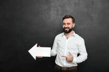 Photo of unshaved man smiling and holding blank speech arrow pointer directing aside, over dark gray wall copy space