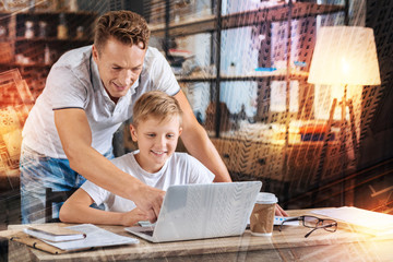 Helpful father. Cheerful positive boy sitting in front of a modern convenient laptop and smiling while his attentive kind responsible father standing behind his back and pointing to the screen