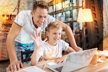 Hello. Cheerful smiling boy waving to the web camera and having a video talk while his attentive kind father standing behind his back and looking happy