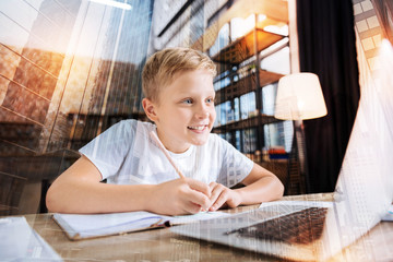 Diligent pupil. Cheerful clever boy sitting in front of a modern laptop and smiling while holding a pen and doing his homework