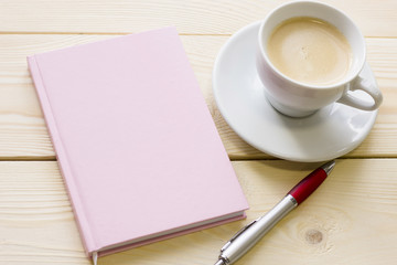 Business Office or Home Workplace Concept with a cup of cappuccino, a dairy in a pink leather cover and a pen on a light wooden table, close up
