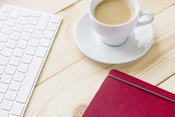 Stylish Business Office or Home Workplace Concept with a cup of cappuccino, a dairy in a red leather cover and a white computer keyboard on a light wooden table, close up