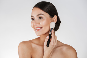 Beauty picture of attractive woman with perfect smile looking aside and applying makeup using brush, isolated over white background