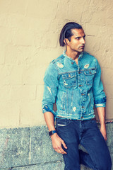 Portrait of Young East Indian American Man in New York. Young handsome man wearing blue fashionable Denim jacket, standing by painted wall, looking away, thinking..