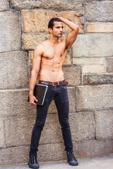 Young East Indian American Man, half naked, showing strong body, wearing black jeans, sneakers, holding laptop computer, standing by rock wall in New York, looking forward..