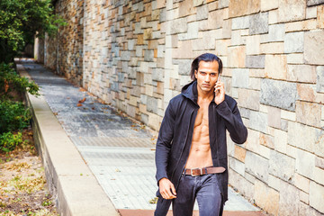 Obraz premium Young East Indian American Man wearing black long hooded outwear, opened, showing half naked strong body, black jeans, talking on cell phone, forcefully walking up along rock wall in New York..