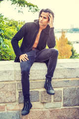 Young Modern East Indian American Man wearing black long hooded outwear, opened, black jeans, sneakers, sitting on stone fence by Hudson Rive in New York, hand touching head, relaxing, thinking...