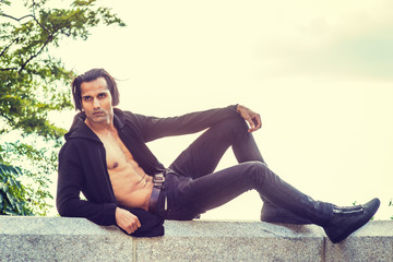 East Indian American Man wearing black hooded outwear, opened, showing half naked body, black jeans, sneakers, sitting on stone fence by Hudson Rive in New York, looking away, relaxing, thinking..