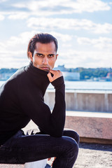 Fototapeta na wymiar Young East Indian American Man wearing black knit pull over sweater, jeans, sitting by river at park in New York, hand holding high collar over chin, intensely looking at you. Fashion style..