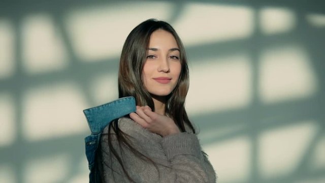 Stylish young sexy brunette woman holds denim jeans jacket before white wall with shadows, indoor slowmotion