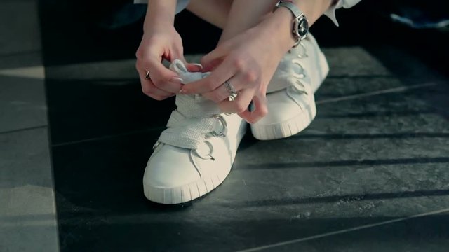 Young stylish girl sitting on the floor wears jeans and white choes, binding the laces, indoor slowmotion