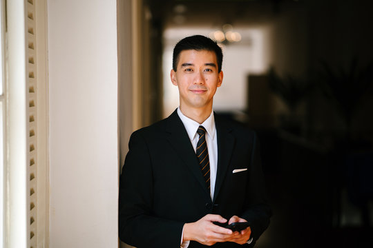 Portrait of young handsome man standing in a well lit corridor. He is wearing formal business attire and is smiling up from his phone.