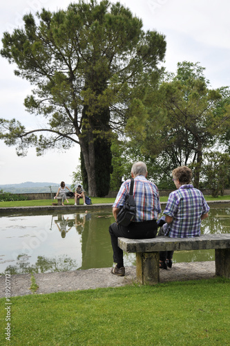 Romantic Old Couple Sitting On Park Bench By Lake In Front Of