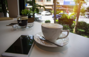 Closeup black coffee cup put on table beside mobile phone and put at the back of blurred cappuccino cup at coffee shop area,blurry light background.