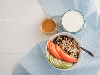 Healthy and tasty breakfast - muesli, slices of persimmon and apple, a glass of fresh milk and maple syrup in a cup on a white table top