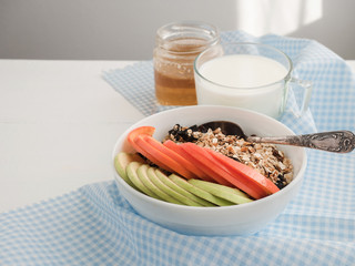 Healthy and tasty breakfast - muesli, slices of persimmon and apple, a glass of fresh milk and maple syrup in a cup on a white table top