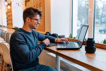 Smiling young man in eyeglasses working on laptop, using graphics tablet and interactive pen display, sitting at the table at cafe with camera and lens. Dressed in sweater and jeans.