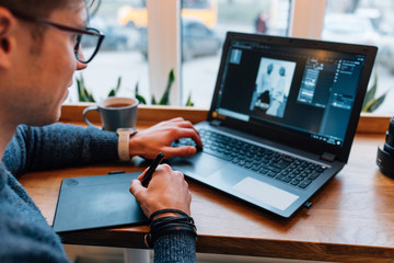 Man is editing photos on laptop, using graphics tablet and interactive pen display, sitting at the table at cafe with cup of tea. Close-up.