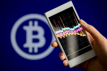 Hand holding smart phone with chart on screen. Blue screen with bitcoin symbol on the background. Crypto-currency concept