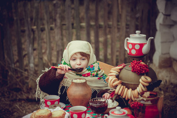 A little girl in an old fur coat and ornament scarf eating porridge with a wooden spoon for the holiday table. Shrovetide Maslenitsa