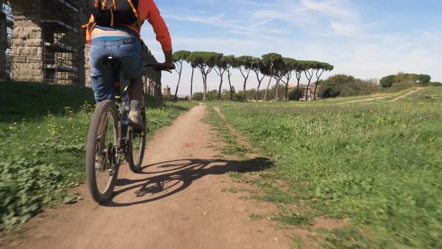 Cyclist riding bike on dirt road leaving dust behind. Young attractive athletic man with orange sportswear and backpack in parco degli acquedotti in Rome close up dolly shot with steadycam
