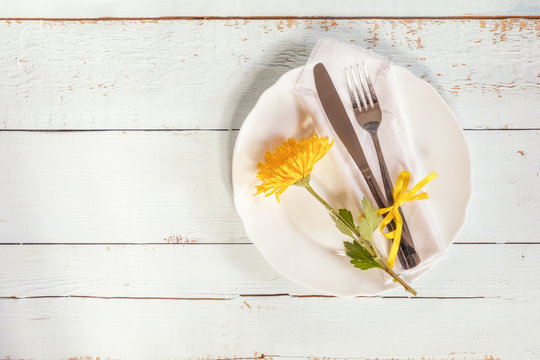 White empty plate, yellow chrysanthemum flowers, napkin, fork and knife tied with a yellow ribbon on light wooden background.