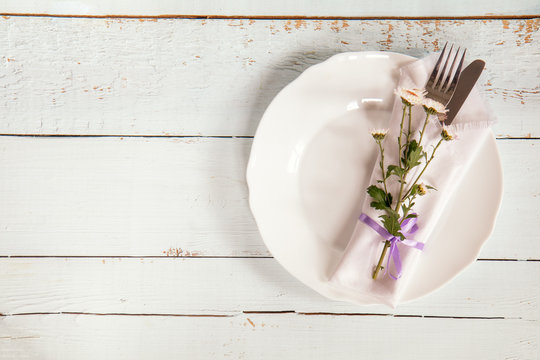 White empty plate, pink chrysanthemum flowers, napkin, fork and knife tied with a violet ribbon on light wooden background.