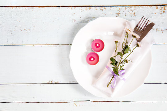 White empty plate, pink chrysanthemum flowers, two pink candles, napkin, fork and knife tied with a violet ribbon on light wooden background.