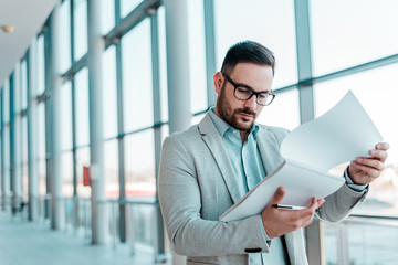 Serious young business man is reading documents while standing near window.