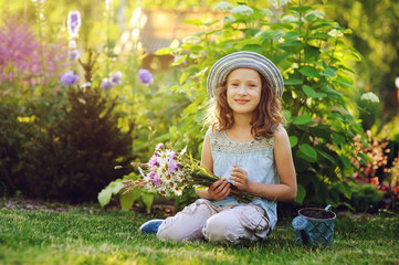 happy child girl playing little gardener in summer, wearing funny hat and holding bouquet of flowers. Sitting on green lawn, enjoying summertime vacations outdoor