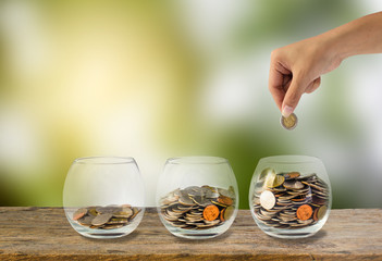 Investment concept. Hand holding putting coins into three step clear glass bottle on wooden table with green blurred background and light. Conceptual saving money for growing business and future