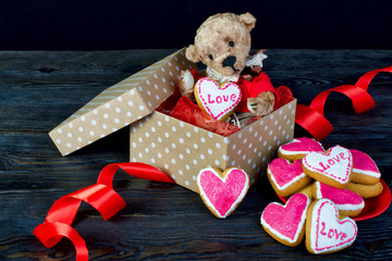 Nice teddy bear sitting in a gift box with a heart. In the hands of a cookie with the inscription "love". Near lie the hill is a cookie-heart. Concept Valentine's Day or a gift for a loved one. 