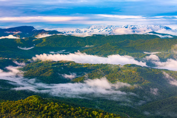 Obraz premium The valley is covered with a spring green forest with rare clouds and fog on the back of the majestic great snowy peaks. Panorama of the Caucasian mountains from the observation tower on Mount Akhun.
