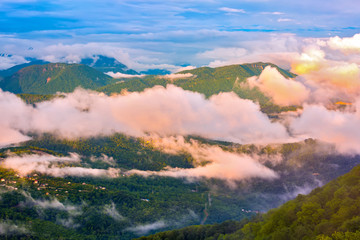 The mountain valley is covered with a spring green forest with rare clouds and fog on the background of a cloudy sky. Panorama of the Caucasian mountains from the observation tower on Mount Akhun.