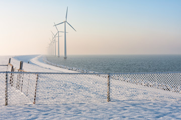 Dutch dike in winter with snow, fence and long row of wind turbines offshore in the water...