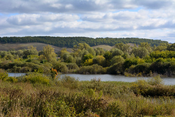 Landscape with meadow, trees, forest on hills and river under cloudy sky. Early autumn in Russia.