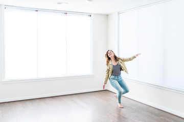One young happy smiling woman jumping up in empty modern new room with hardwood floors and large...