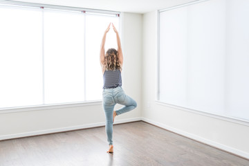 Young woman in tree yoga pose standing on one leg in empty modern new room, hardwood floors, large sunny windows in apartment, house