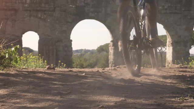 Cyclist riding bike on dirt road leaving dust behind. Young attractive athletic man with orange sportswear and backpack in parco degli acquedotti in Rome close up dolly shot with steadycam