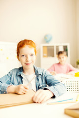 Pretty little schoolgirl with crayon and notepad attending lesson of drawing