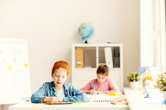 Two little classmates sitting by individual desks and carrying out teacher task
