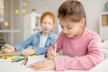 Diligent girl with crayon drawing at lesson while her classmate looking at her notepad