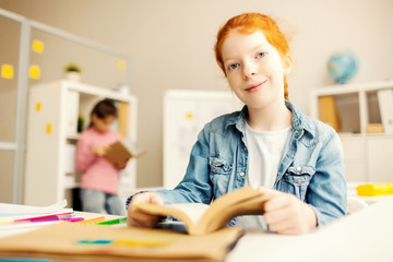 Cute schoolgirl with book looking at camera while sitting in library after lessons