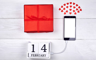 Top view of mobile smart phone with white blank screen, red gift box, hearts and wooden block calendar on wooden background, copy space. Flat lay, top view. Valentines day application mockup template