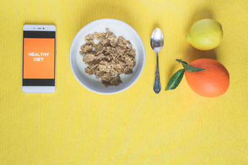Top view of a white bowl of cereal, orange, lemon, and a mobile phone with written Healthy Diet on the screen on bright yellow background with copy space. Fitness, healthy diet or breakfast concept