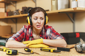 Bewildered perplexed upset young woman in plaid shirt, gray T-shirt, noise insulated headphones, yellow gloves working in carpentry workshop at wooden table place with piece of wood, different tools.