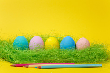 Row of five colorful pastel monophonic painted Easter eggs, pencils in green grass on yellow background for card or postcard. Happy Easter concept. Copy space for advertisement. With place for text.