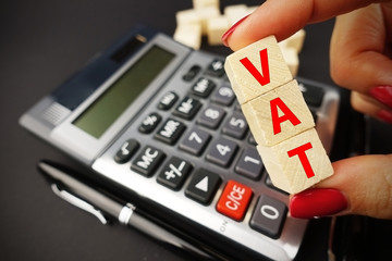 VAT calculation concept with woman hand holding vat word on wooden cubes above calculator keyboard on dark background
