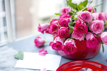 A bouquet of fresh pink roses, in a pan of red cast iron, on a gray stone background. Near the window. White envelope. Free place to write text or postcard.